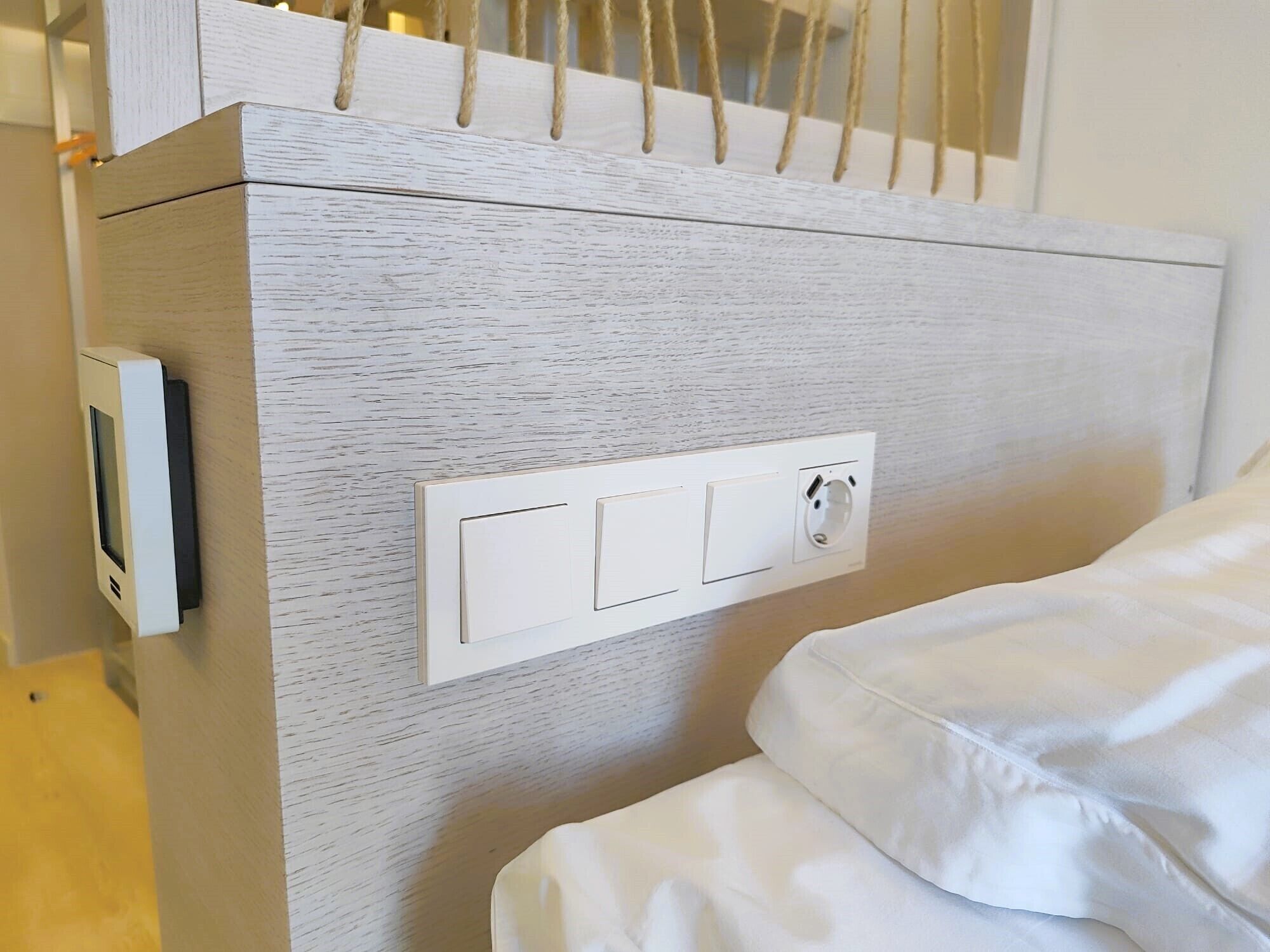 Ein weißes Bett mit Holzkopfseitenverkleidung, einem Nachttisch und einer an der Wand montierten Steuerung.