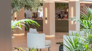 A modern lobby with a seating area featuring two white armchairs and a coffee table. There are indoor plants and a hanging plant above the seating area. The background includes a bar area with a counter and shelves. - Valentin Somni Hotels & Suites – Adults Only +16 (Calvia)