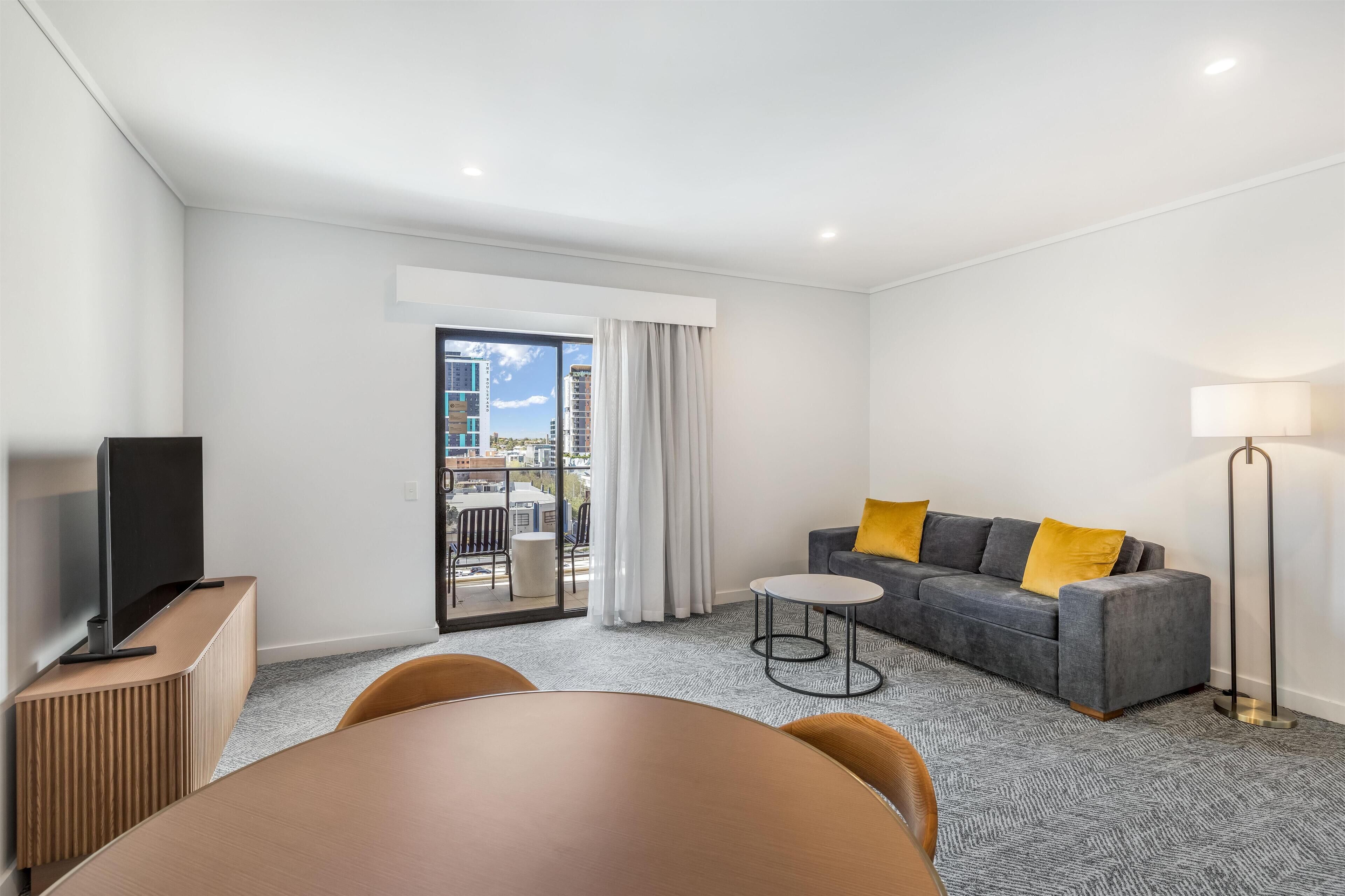A modern hotel room with a flat-screen TV, a grey sofa with yellow cushions, a small round table, and a floor lamp.