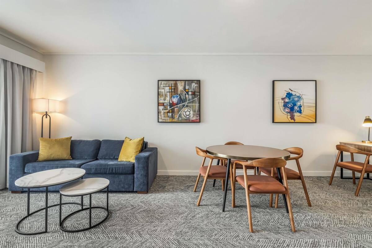 A modern hotel room with a blue sofa, a round dining table with wooden chairs, and two small side tables. There are two framed artworks on the wall, a floor lamp, and a window with curtains.