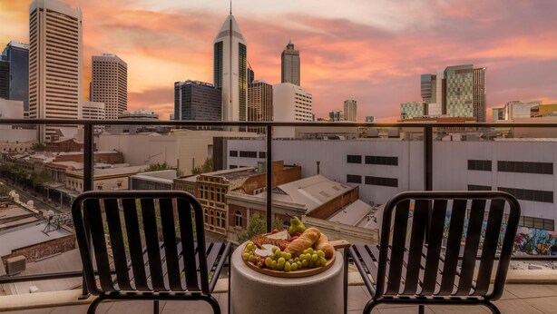 Un balcon avec deux chaises et une table sur laquelle est disposée une assiette de fruits, offrant une vue sur un paysage urbain avec des bâtiments modernes et un ciel au coucher du soleil.