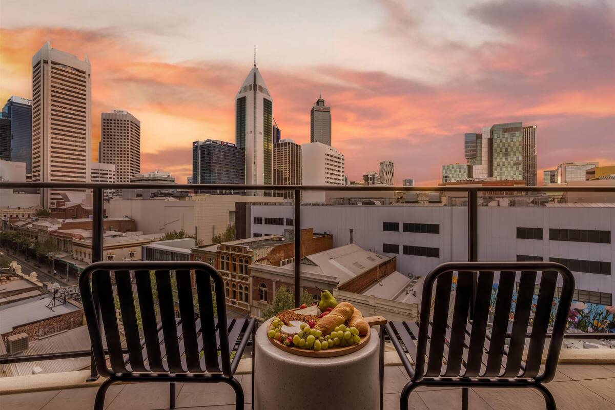 A balcony with two chairs and a table with a fruit platter, overlooking a cityscape with modern buildings and a sunset sky.