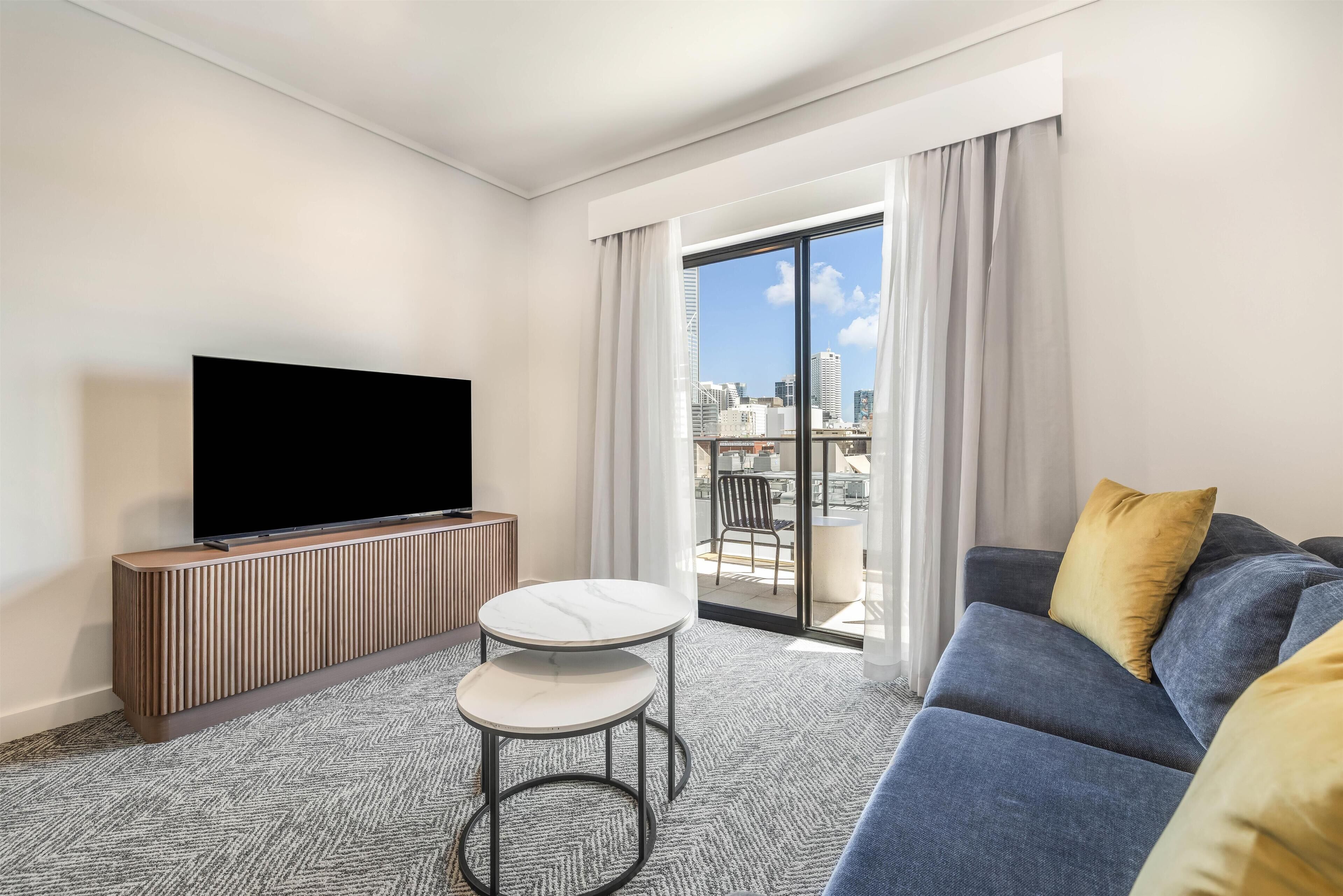 A modern hotel room with a flat-screen TV on a wooden console, a grey sofa with yellow cushions, a small round table, and a city view through large sliding doors.