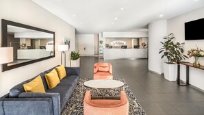 Un hall d’hôtel moderne avec un comptoir d’accueil, un canapé avec des coussins jaunes, une table basse ronde et un grand miroir.
