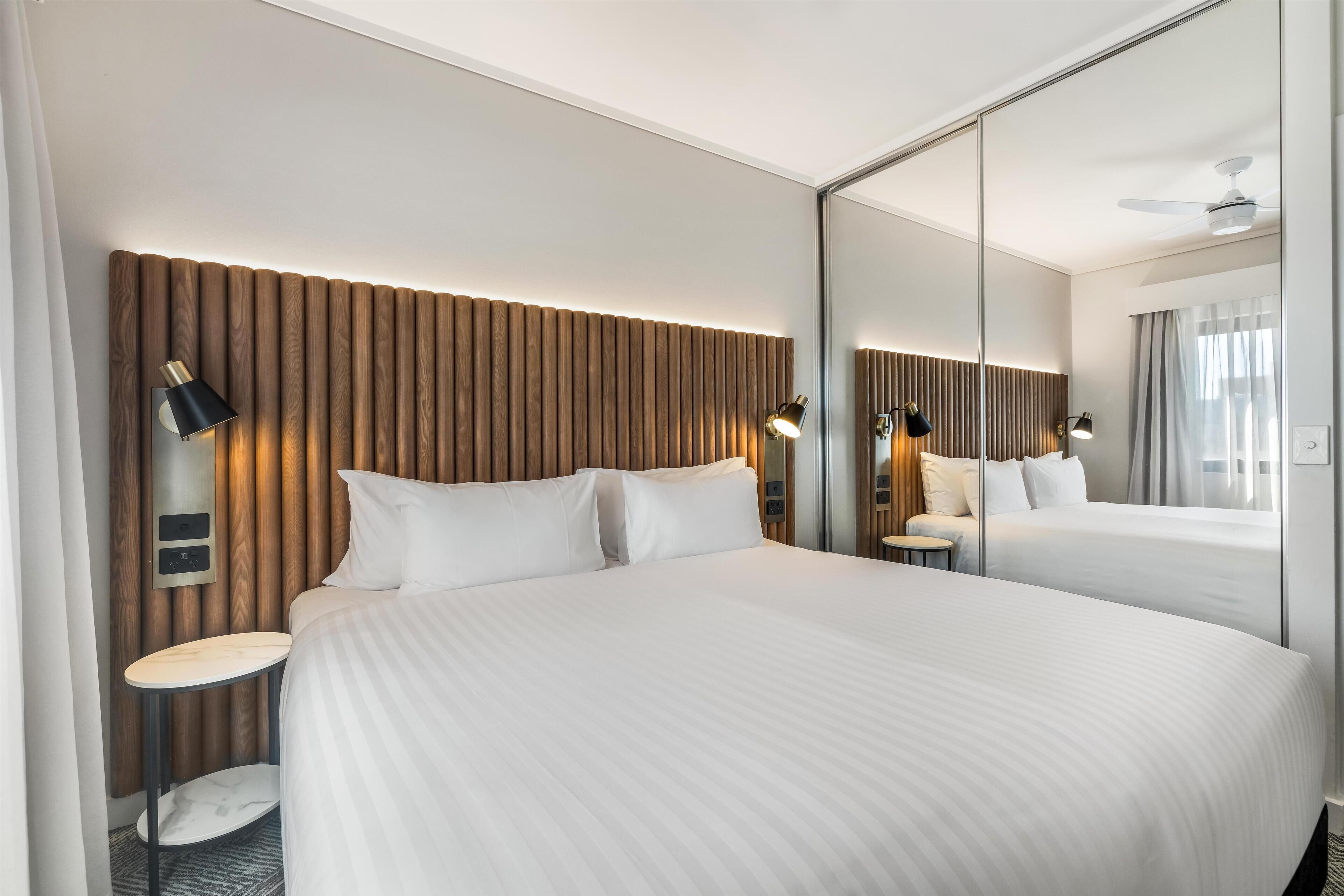 A hotel room with a large bed, a bedside table, a mirror, and a wall-mounted light fixture.