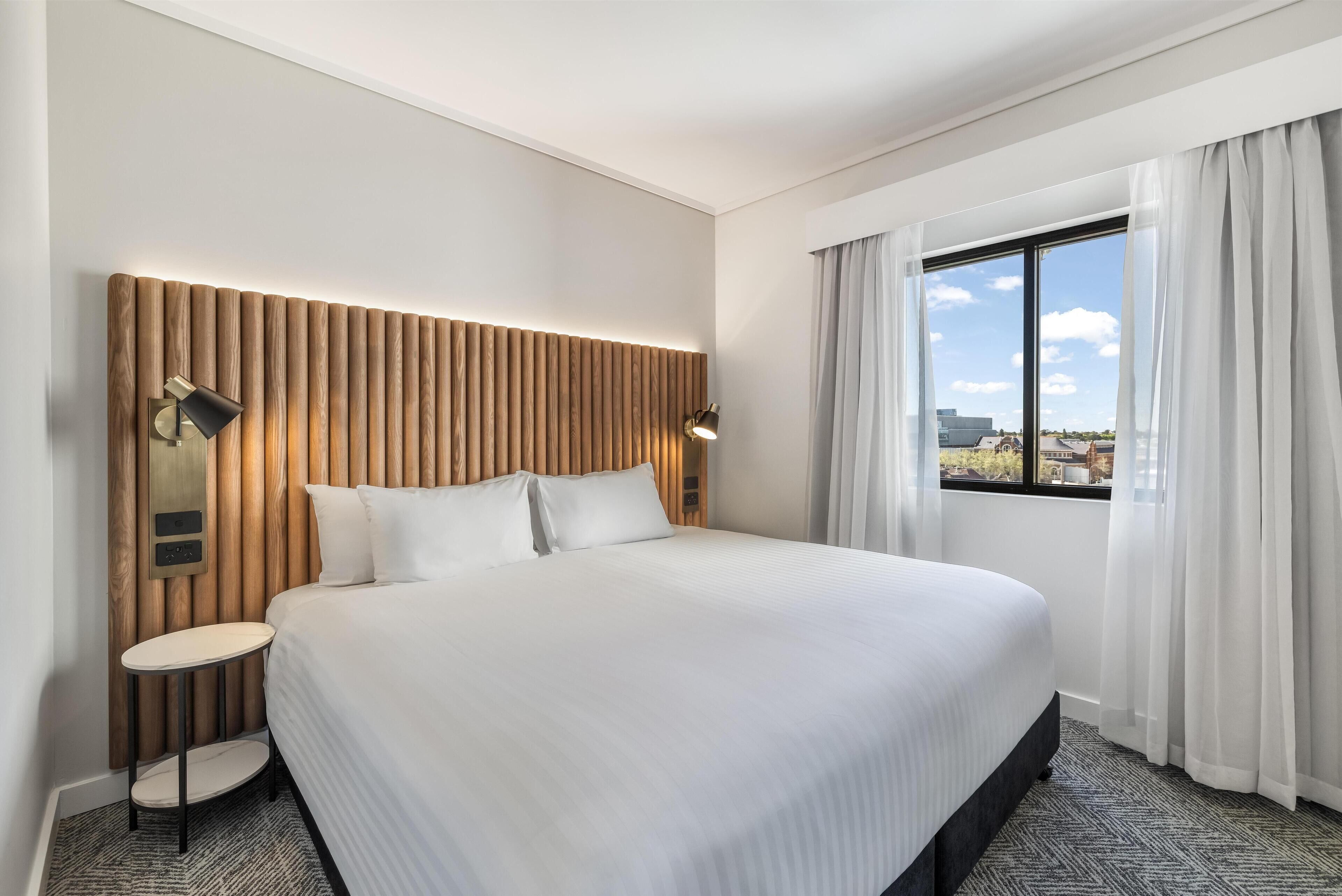 Une chambre d’hôtel moderne avec un grand lit, une tête de lit en bois, une petite table ronde et une fenêtre avec des rideaux.