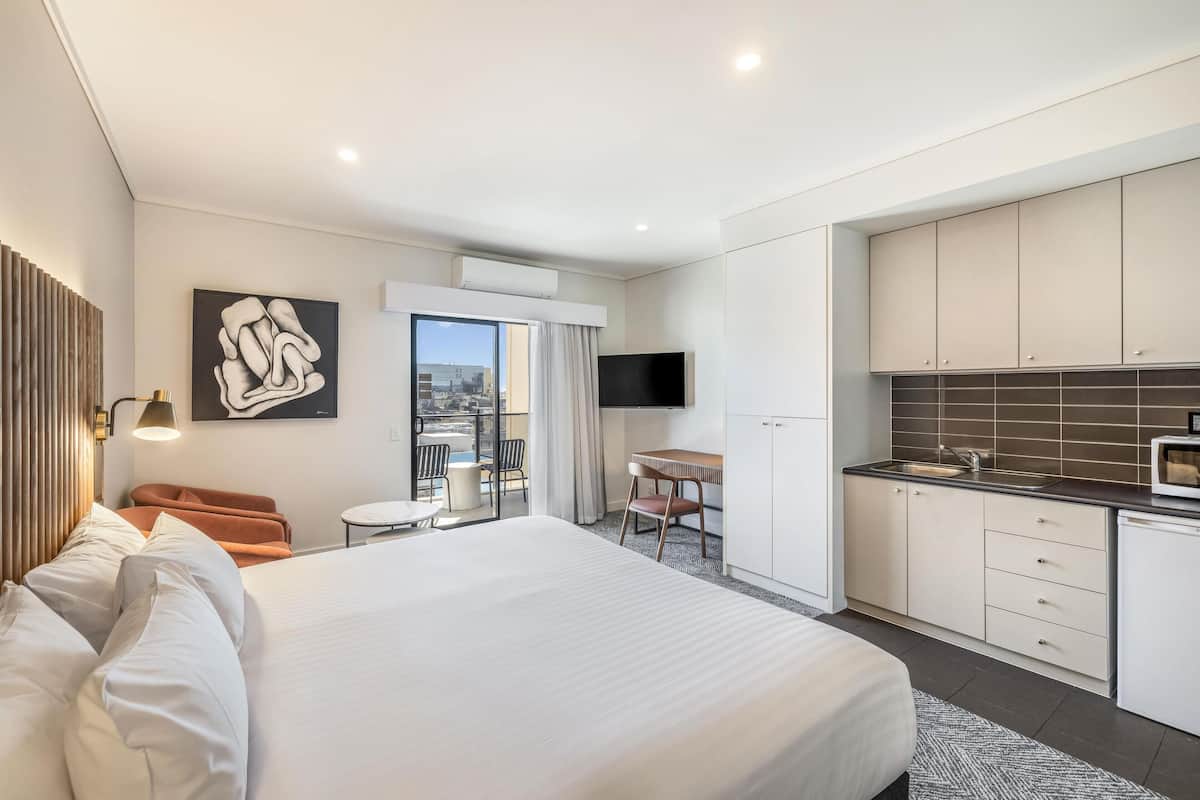 A modern hotel room with a large bed, a desk, a chair, a TV, and a kitchenette.