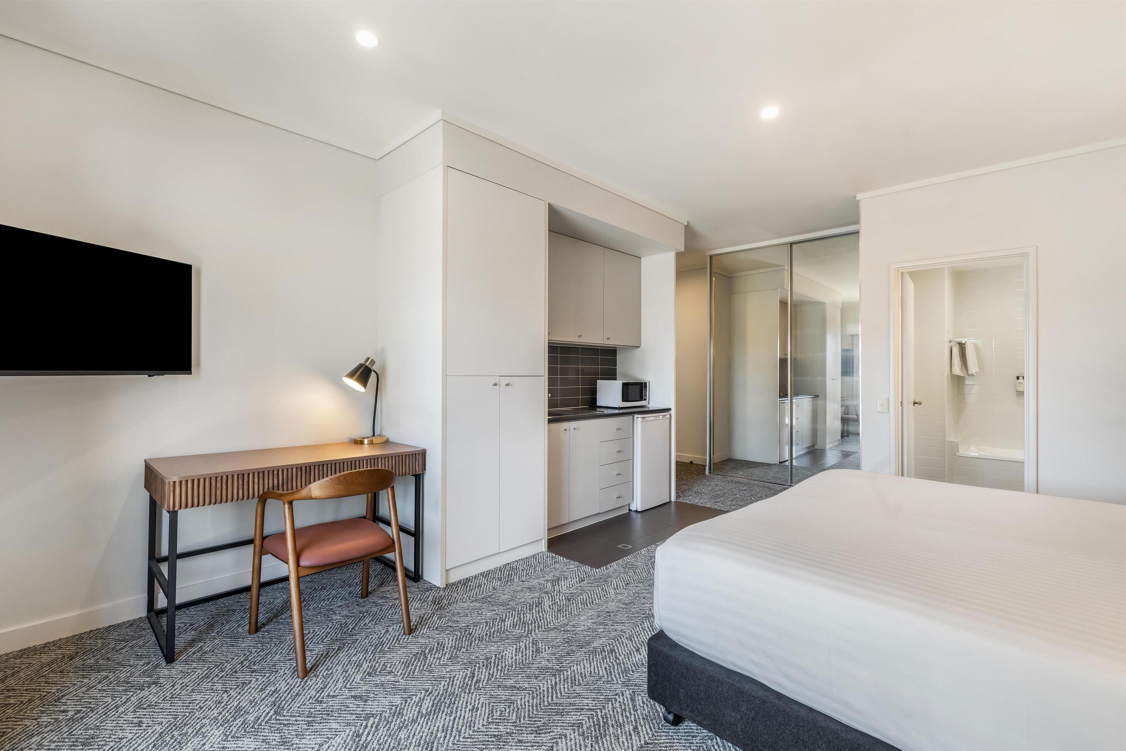 A modern hotel room with a bed, desk, chair, and kitchenette.