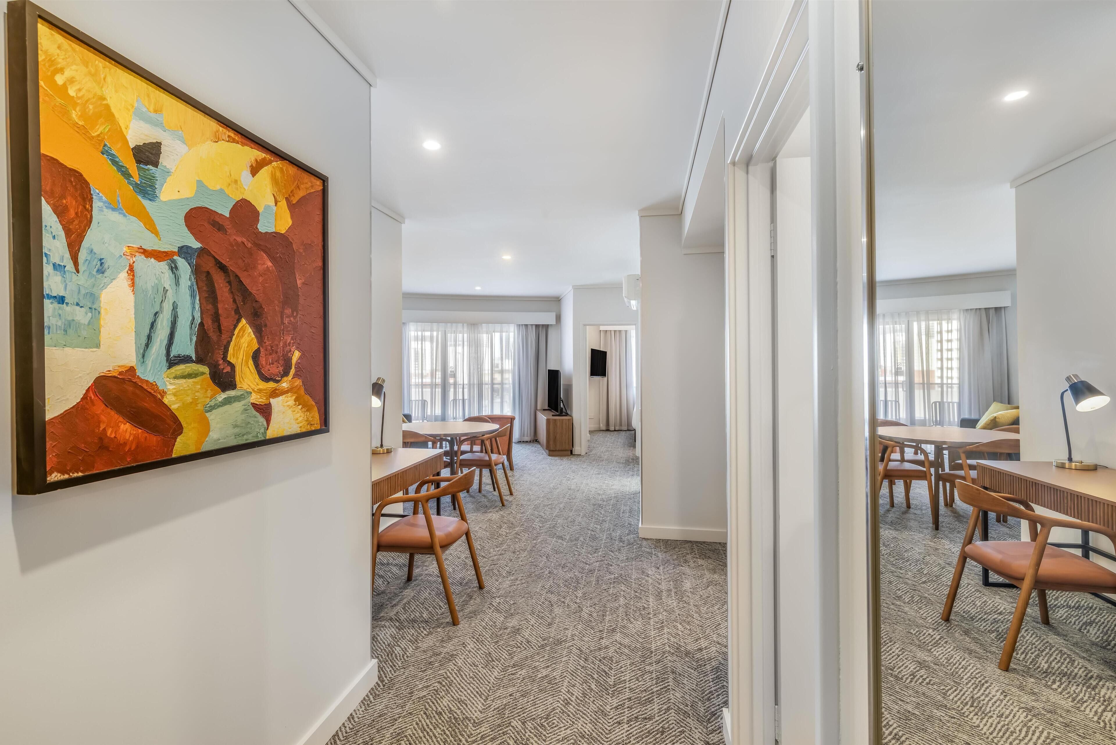A hotel room with a large abstract painting, a dining area with wooden furniture, and a carpeted floor.