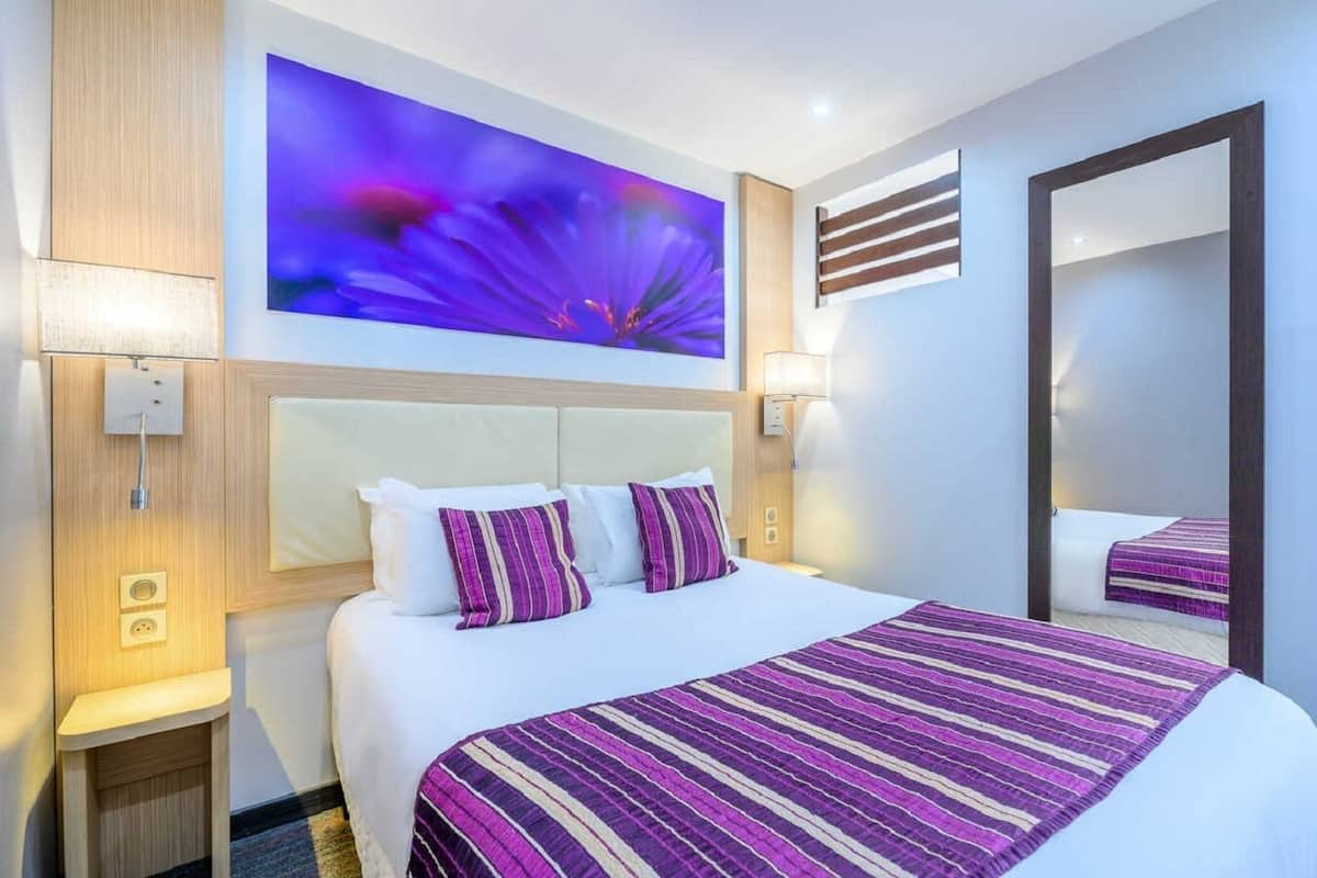 A hotel room with a large bed, a headboard, a wall-mounted artwork, a mirror, and bedside lamps.