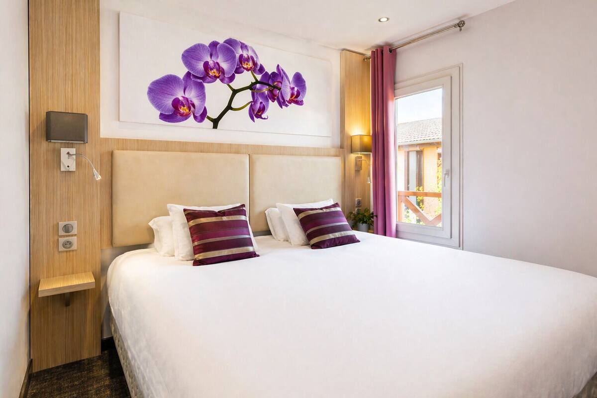 A hotel room with a large bed, a headboard, a wall-mounted lamp, a power outlet, and a window with curtains.