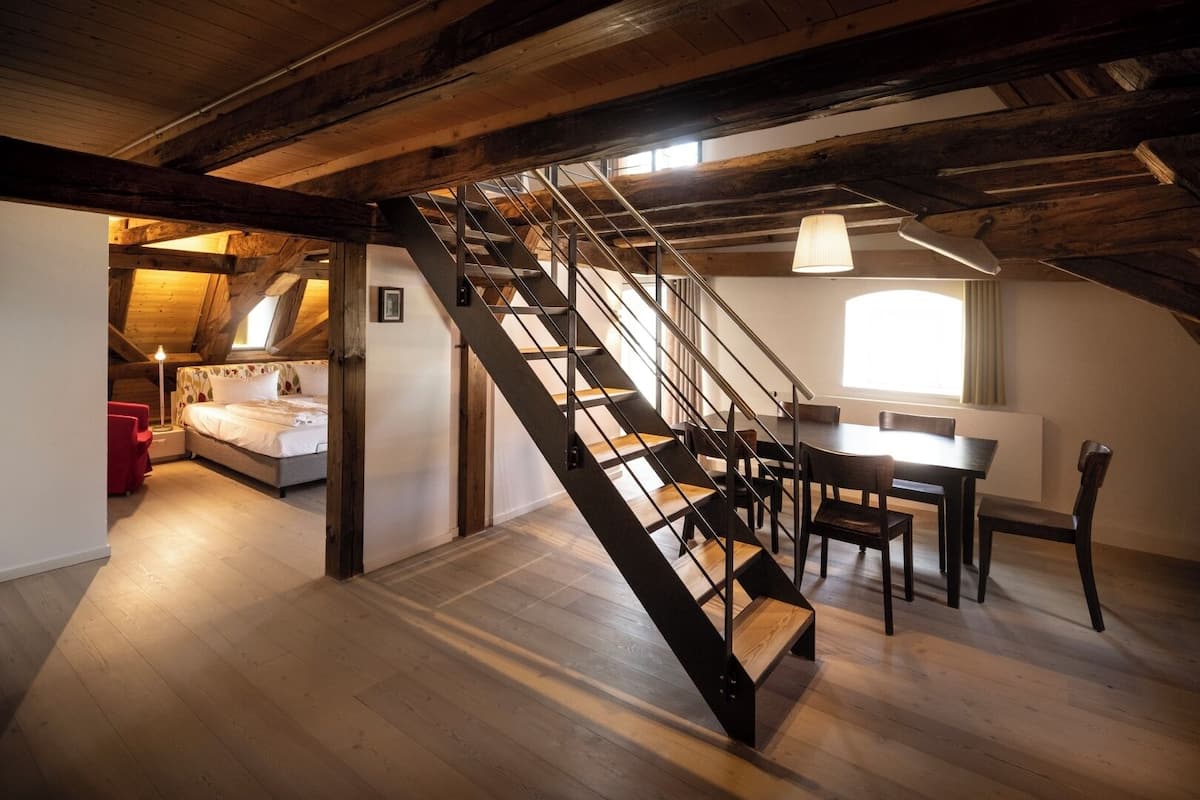 Una habitación estilo loft con escalera de madera, una cama, una mesa de comedor y un ventanal.