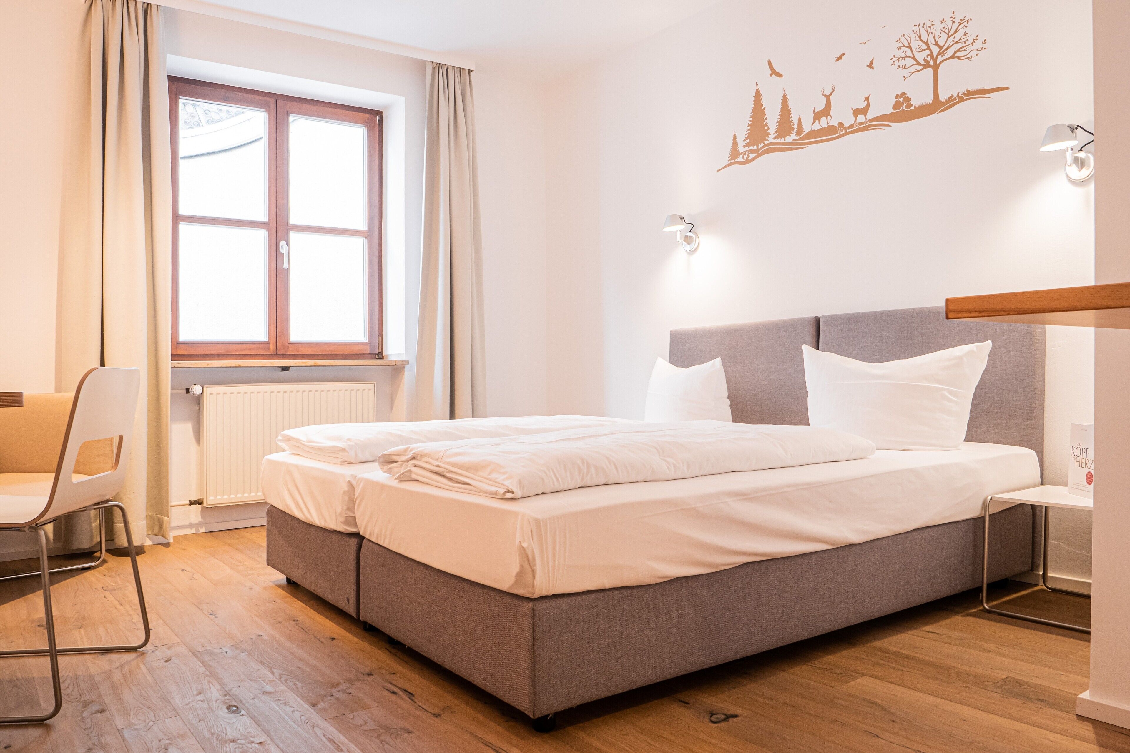 A neatly made bed with white linens, a grey headboard, and a window with beige curtains.