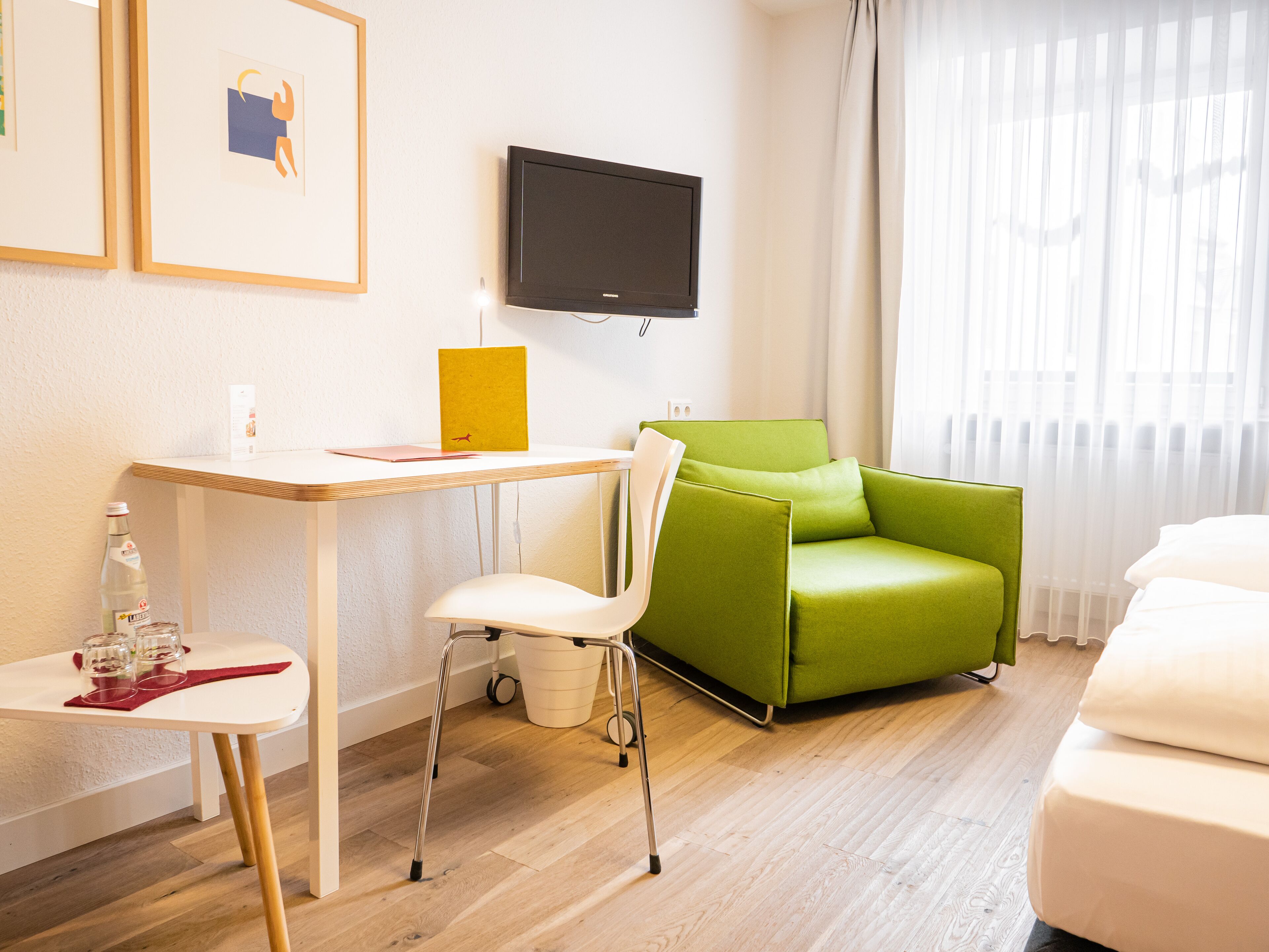 A modern hotel room with a wooden desk, a green armchair, a white sofa, and a flat-screen TV mounted on the wall.