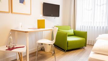 A modern hotel room with a wooden desk, a green armchair, a white sofa, and a flat-screen TV mounted on the wall.