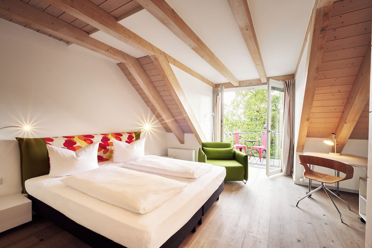 Un dormitorio con una cama grande, una silla verde, un escritorio y un balcón con vistas a la vegetación.