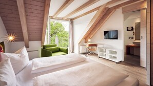 Un dormitorio con techo inclinado, una cama, una silla verde, un escritorio, un televisor y un baño visible a través de una puerta abierta.
