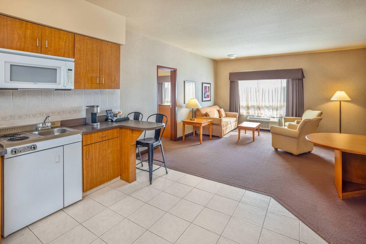 Une chambre d’hôtel dotée d’une kitchenette, d’un coin repas, d’un salon avec un canapé et des fauteuils, et d’une grande fenêtre avec des rideaux.