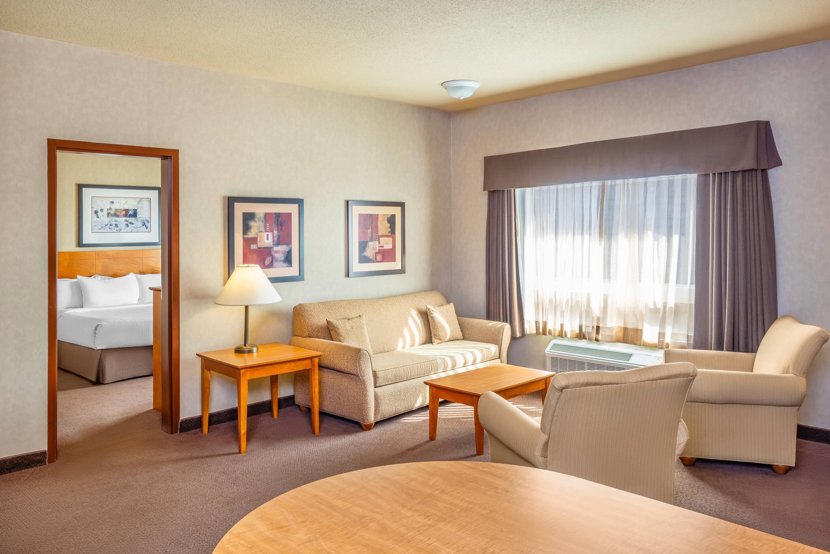Habitación de hotel con sofá, dos sillones, una mesita y cama. En la pared hay cuadros enmarcados y una ventana con cortinas.