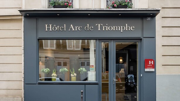 The exterior of Hotel Arc de Triomphe with a large glass entrance, a sign indicating a handicap accessible entrance, and a small balcony with potted plants.