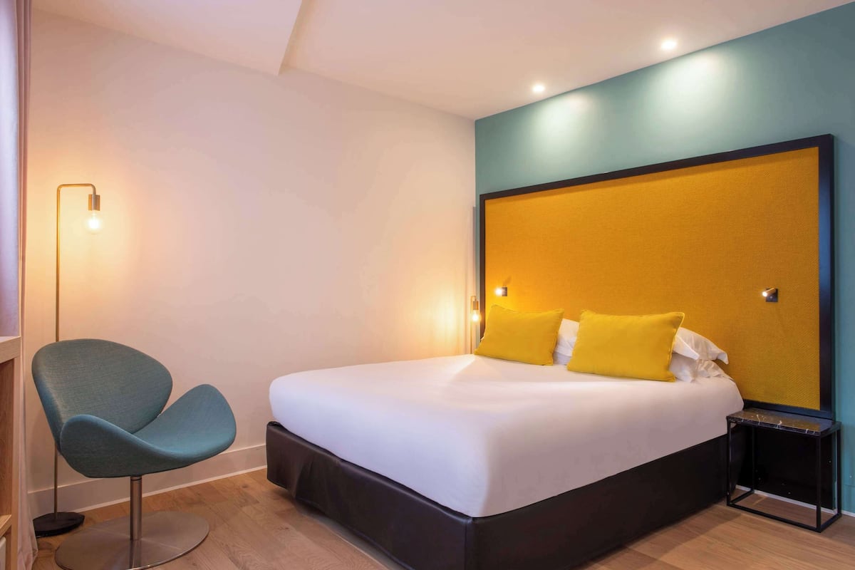 A modern hotel room with a large bed, a single chair, a bedside table, and a floor lamp.