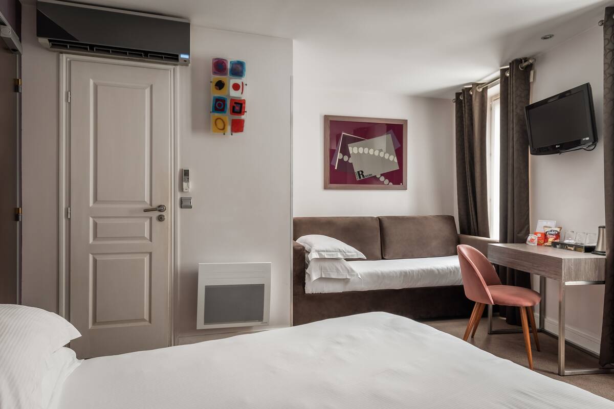 A hotel room with a bed, a desk, a chair, a TV, a door, and a wall with colorful shapes.
