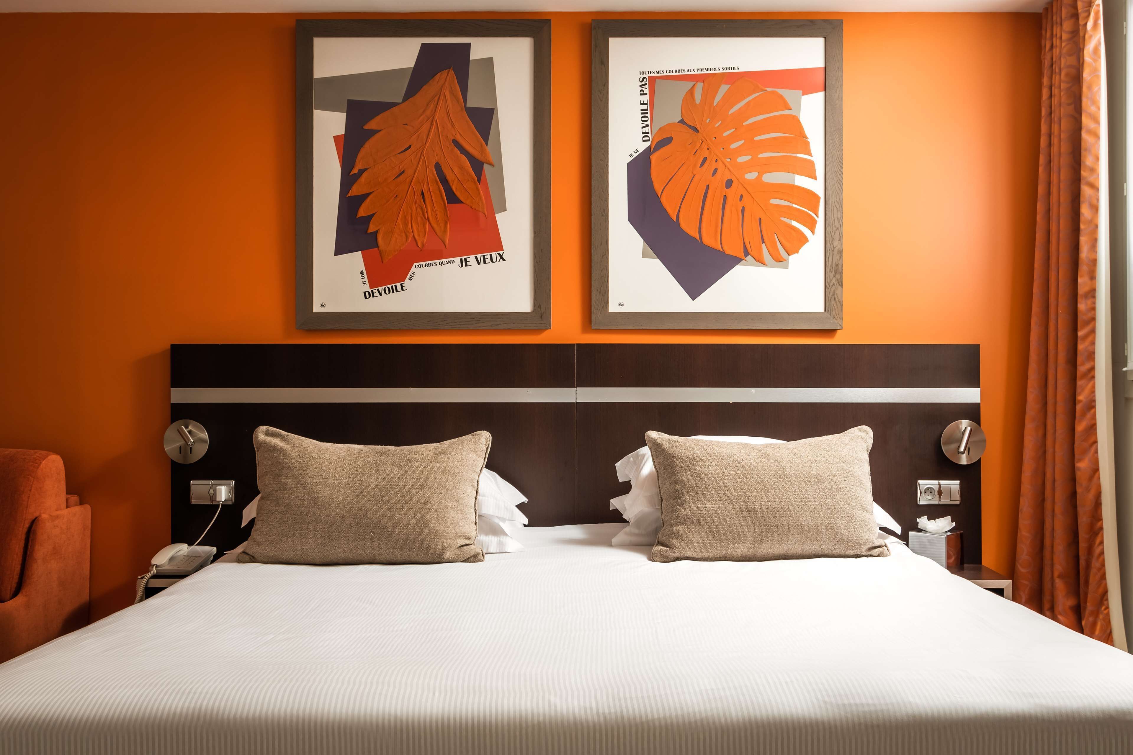 A hotel room with a large bed, two pillows, a nightstand with a phone, and two framed artworks on the wall.
