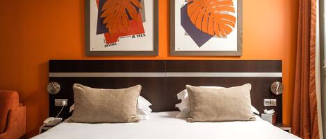 A hotel room with a large bed, two pillows, a nightstand with a phone, and two framed artworks on the wall.