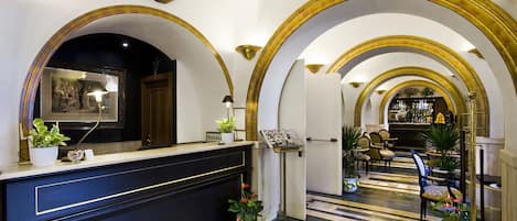A hotel lobby with a reception desk, framed artwork, potted plants, and a bar area in the background.