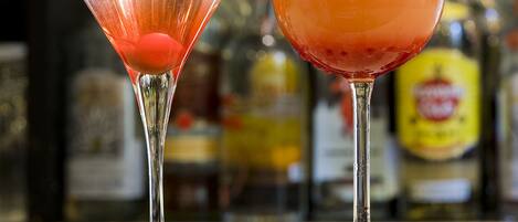Two cocktails on a bar counter with a blurred background of bottles.