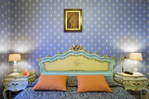 A bedroom with a patterned wallpaper, a decorative headboard, two bedside tables with lamps, and two pillows.