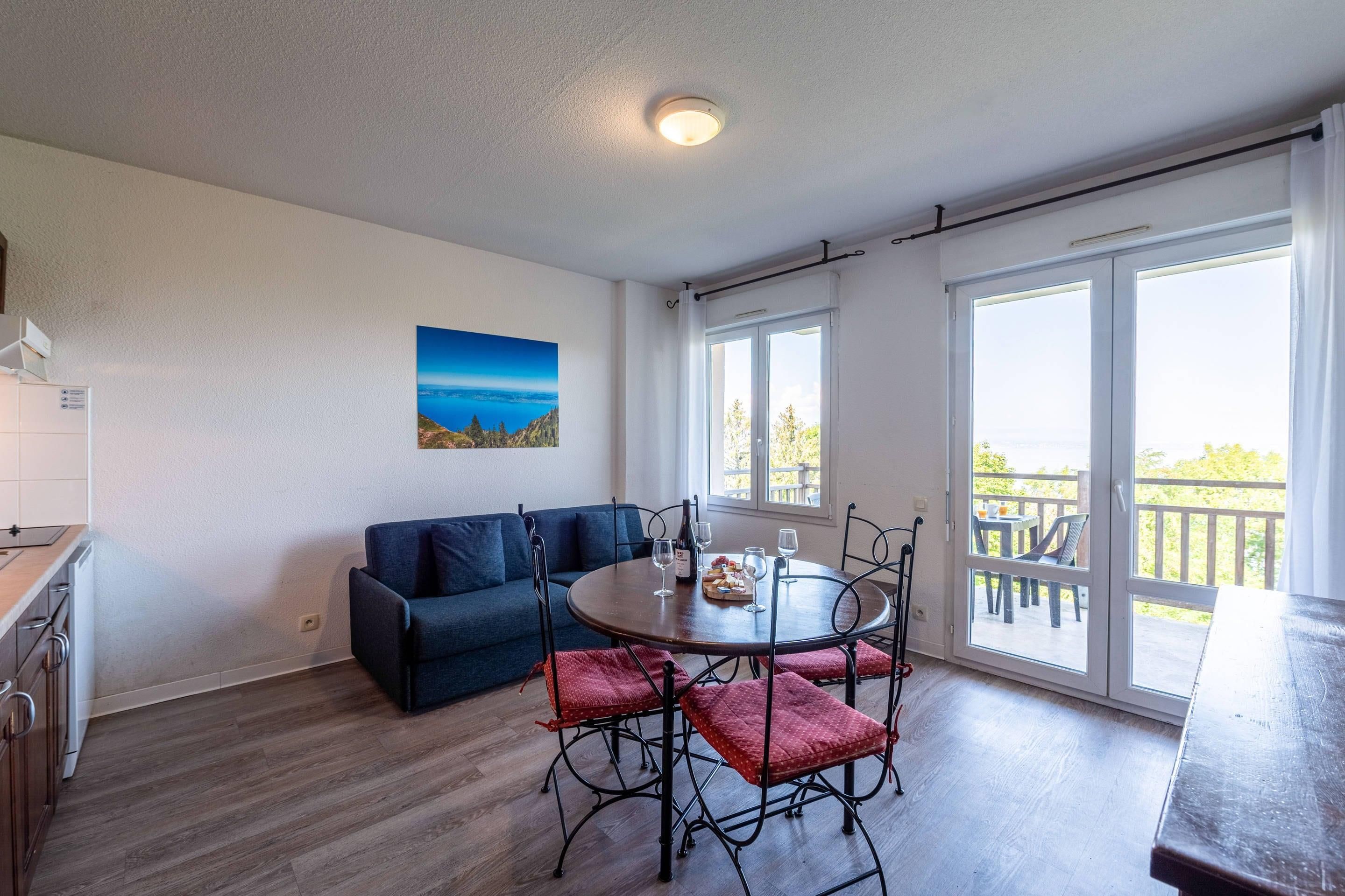 Eine moderne Küche mit Backofen, ein Essbereich mit rundem Tisch und rot gepolsterten Stühlen, ein blaues Sofa und ein großes Fenster mit Blick ins Freie.