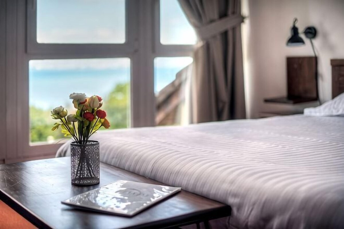 A bed with a view of the sea, a bedside table with a vase of flowers, and a lamp on the wall.