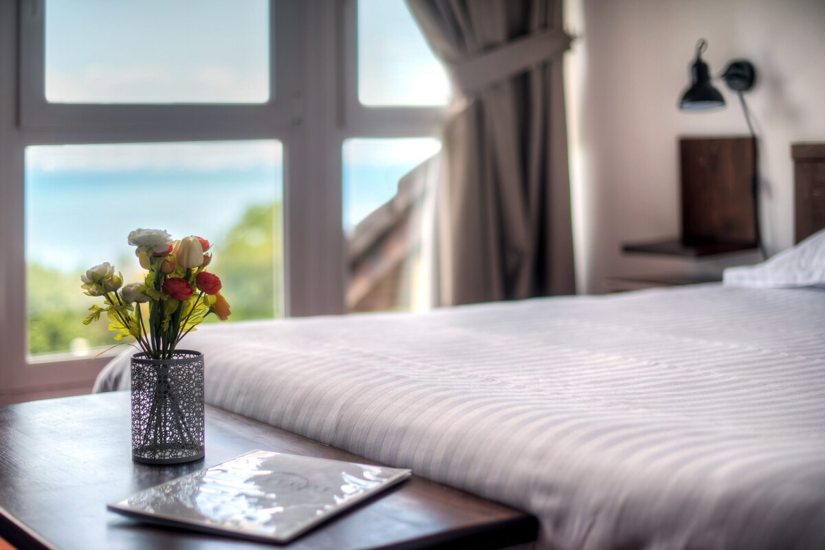A bed with a view of the outdoors, a bedside table with a vase of flowers, and a lamp on the wall.
