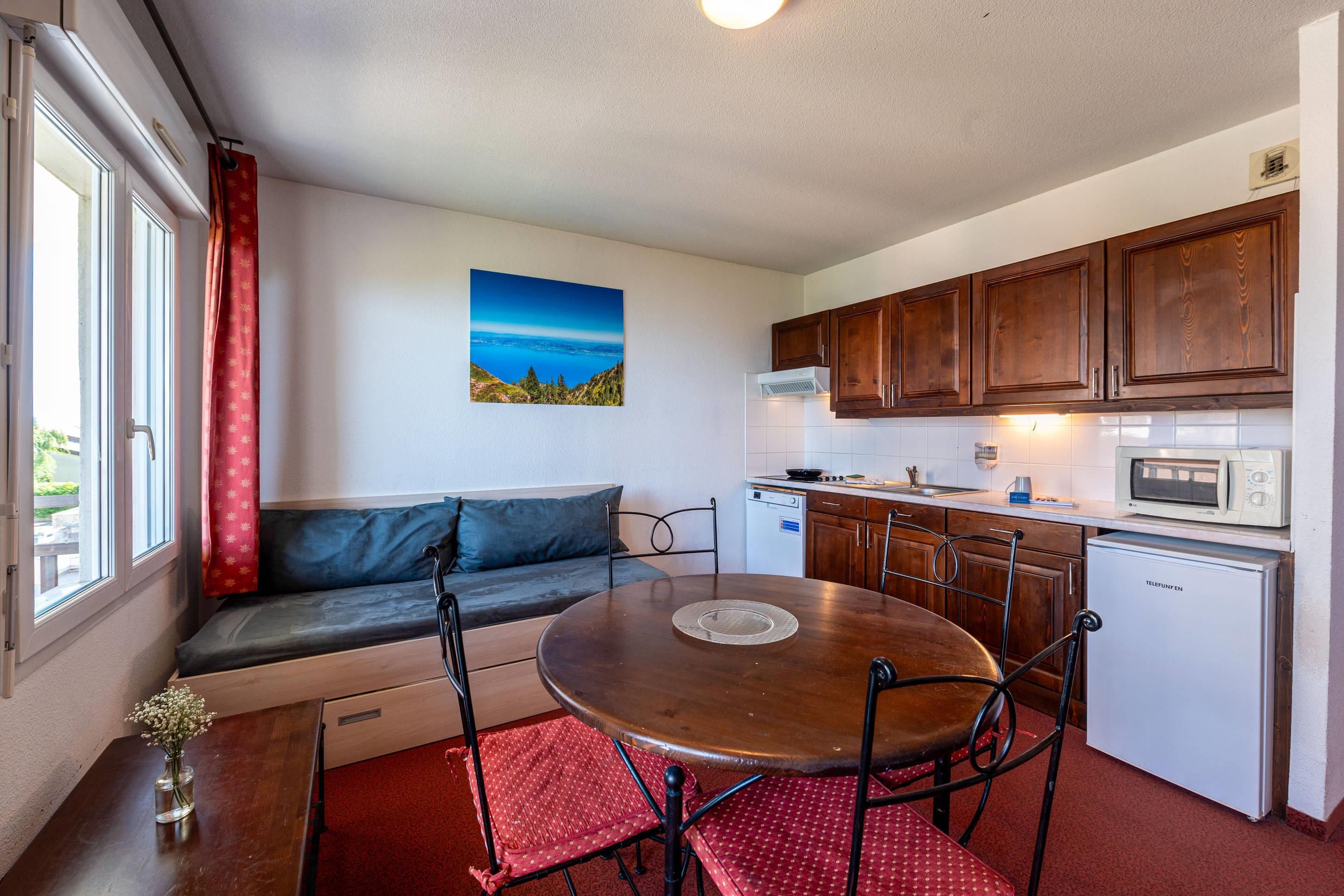 A compact kitchen with wooden cabinets, a microwave, and a small refrigerator. There is a dining table with chairs and a sofa with a view of the outdoors.