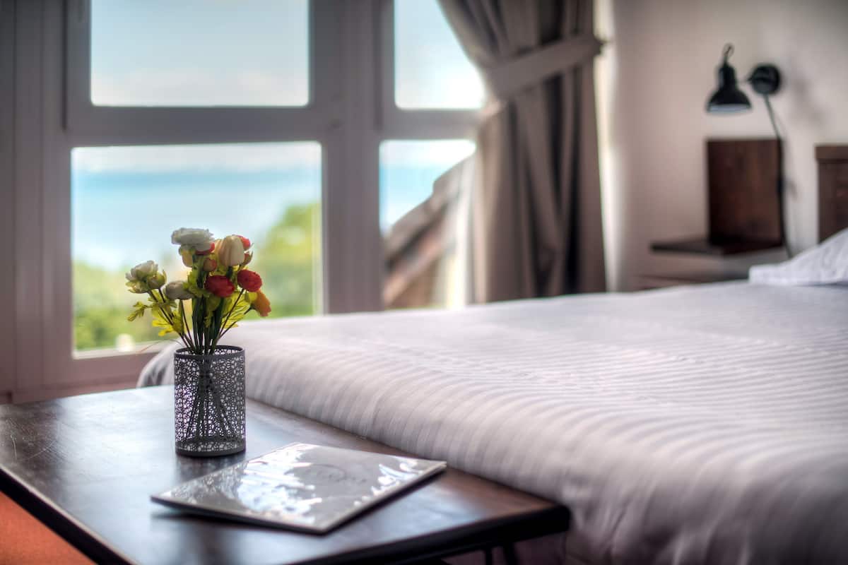 A bed with a white comforter, a vase with flowers on a bedside table, and a view of the outdoors through a window.