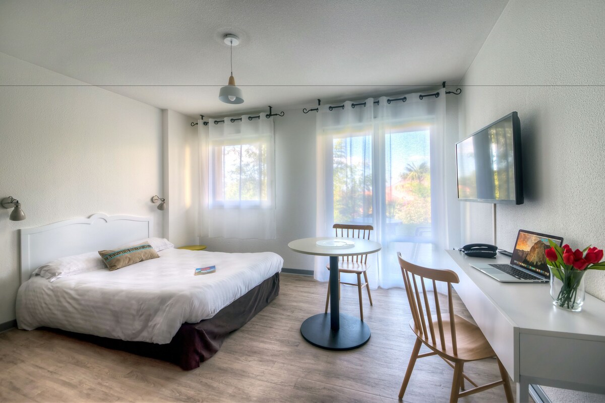 A bedroom with a bed, a desk with a laptop, a chair, a small table, and a TV mounted on the wall.