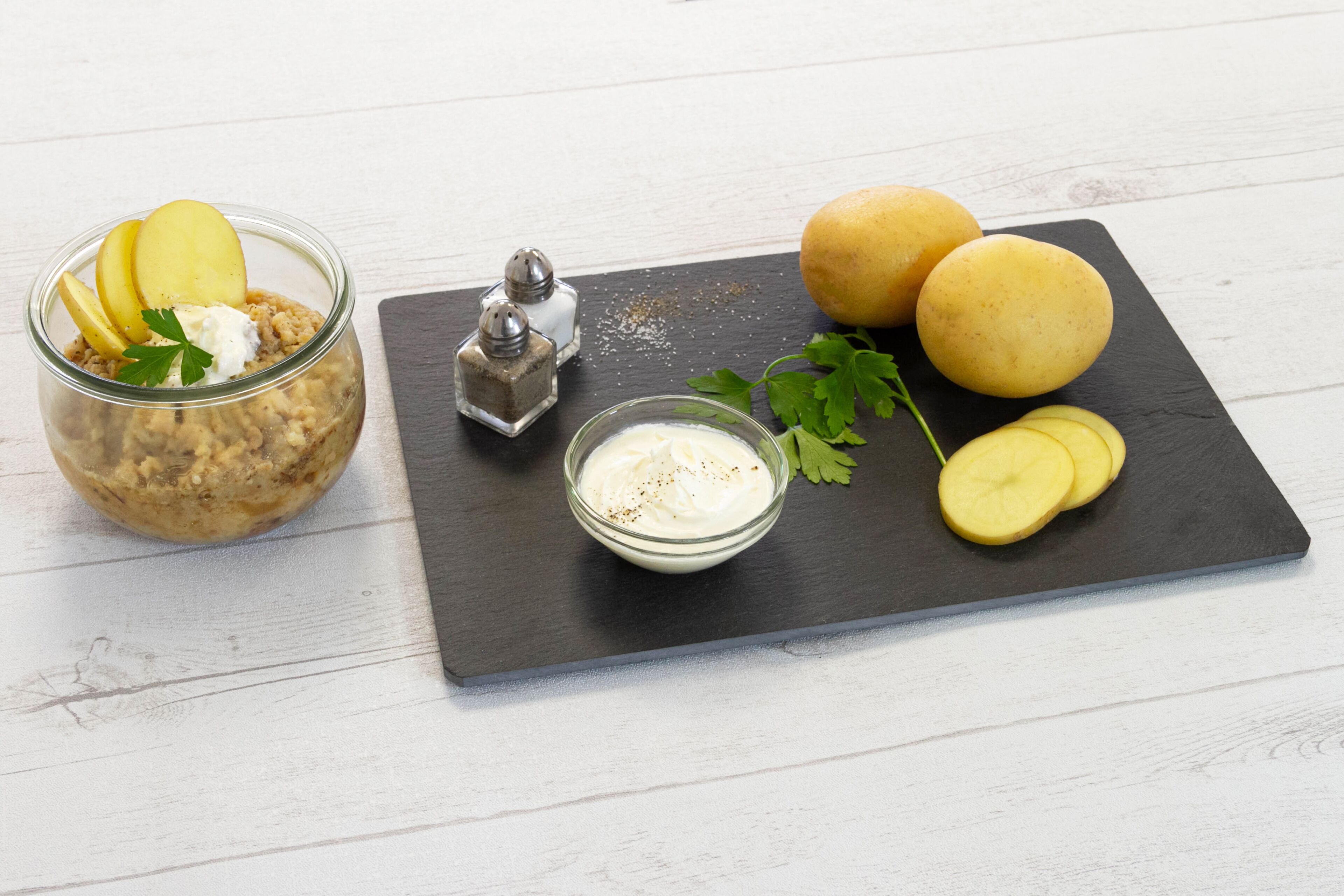 A food arrangement with a jar, two potatoes, a bowl of sauce, and a salt shaker on a slate board.