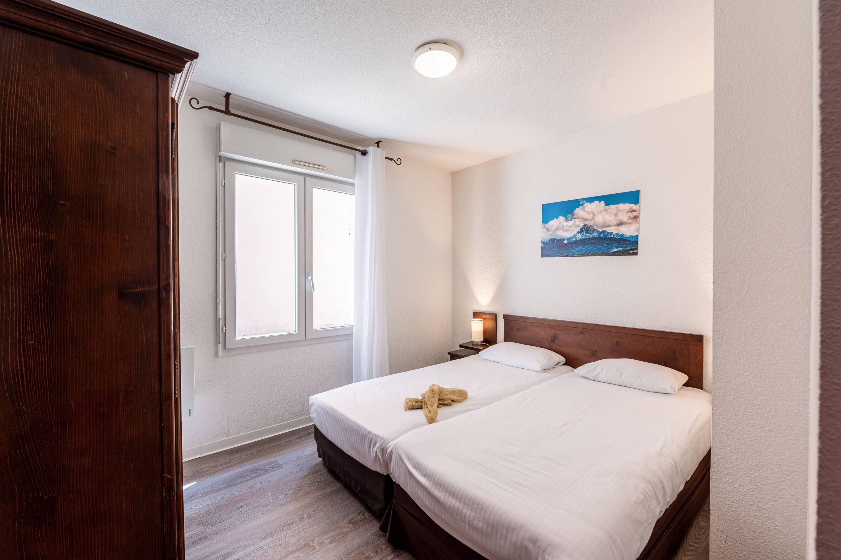 Habitación de hotel con dos camas, un armario de madera, una ventana con cortinas, una mesita de noche con lámpara y un cuadro en la pared.