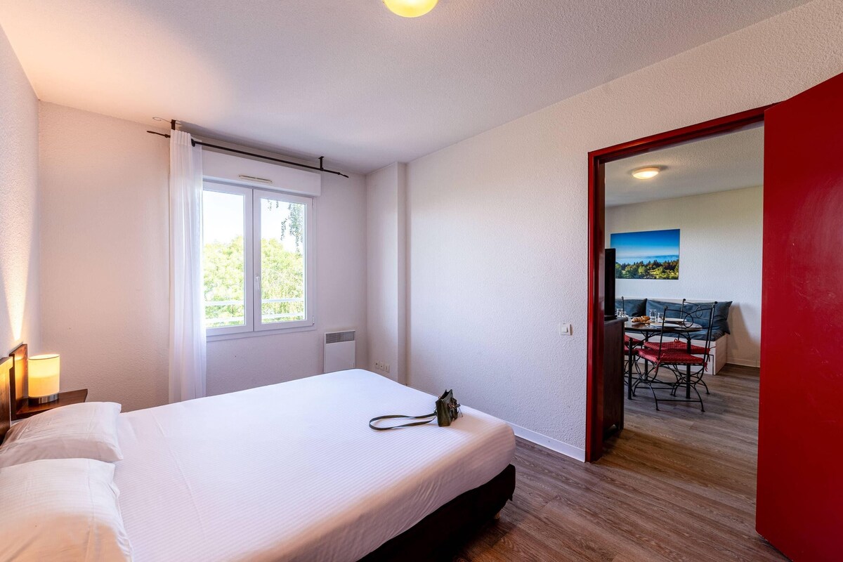 A bedroom with a bed, a window, a door leading to a dining area, and a lamp on a nightstand.