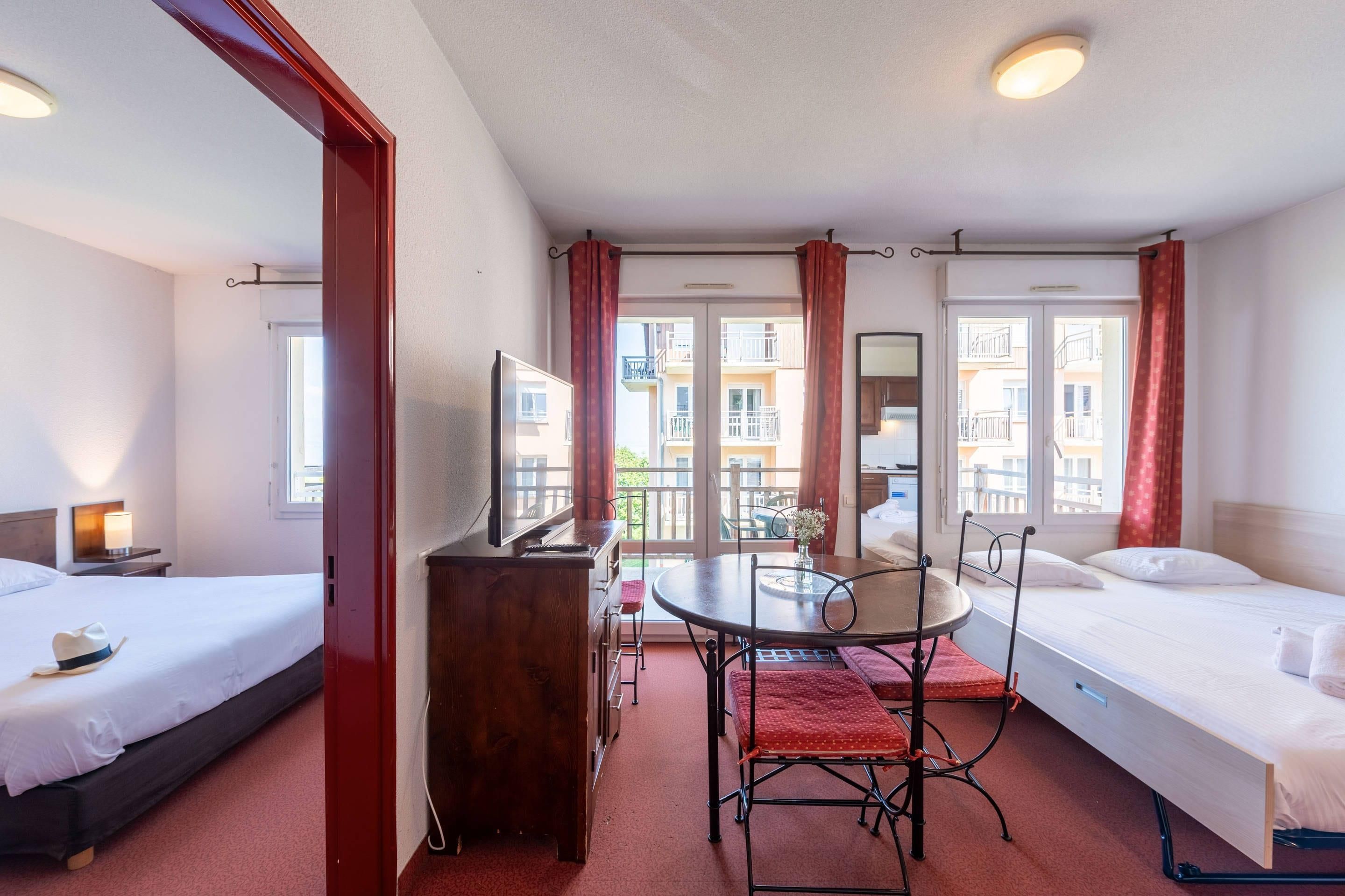 Et hotelværelse med to senge, et lille bord, et fjernsyn og en balkon med røde gardiner.