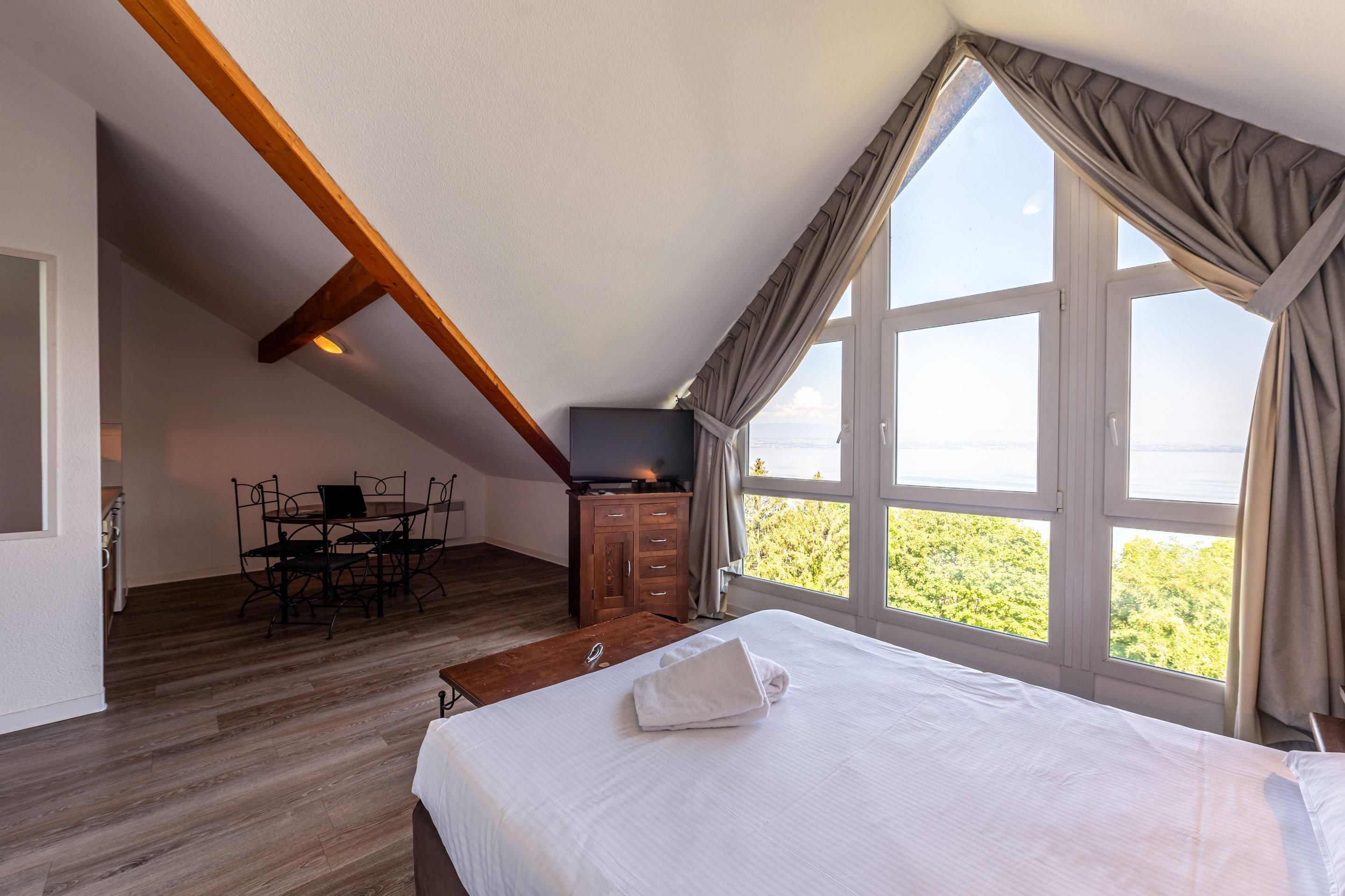 Une chambre avec une grande fenêtre, un lit, une table en bois et un téléviseur.