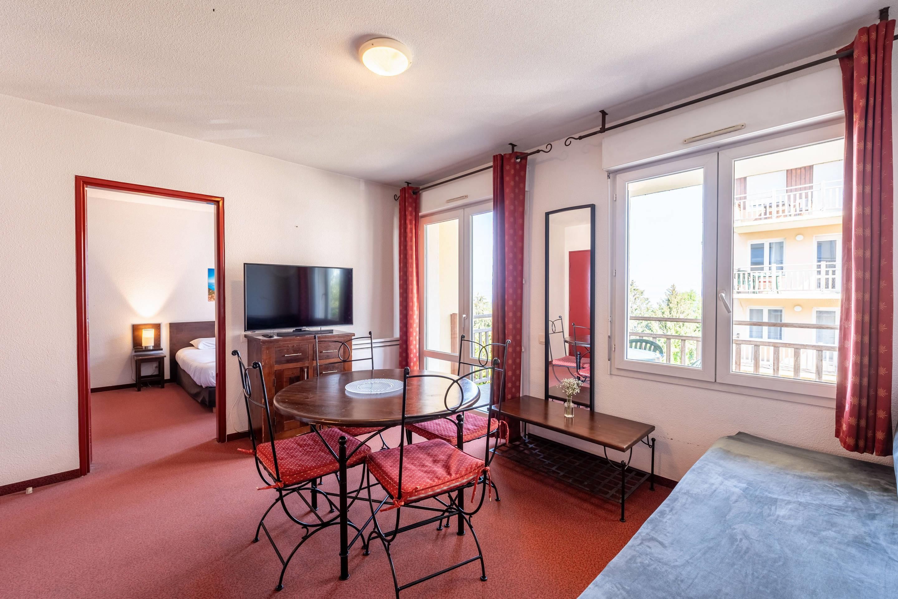 A hotel room with a dining table, chairs, a TV, a bed, and large windows with curtains.