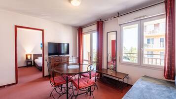 A hotel room with a dining table, chairs, a TV, a bed, and large windows with curtains.