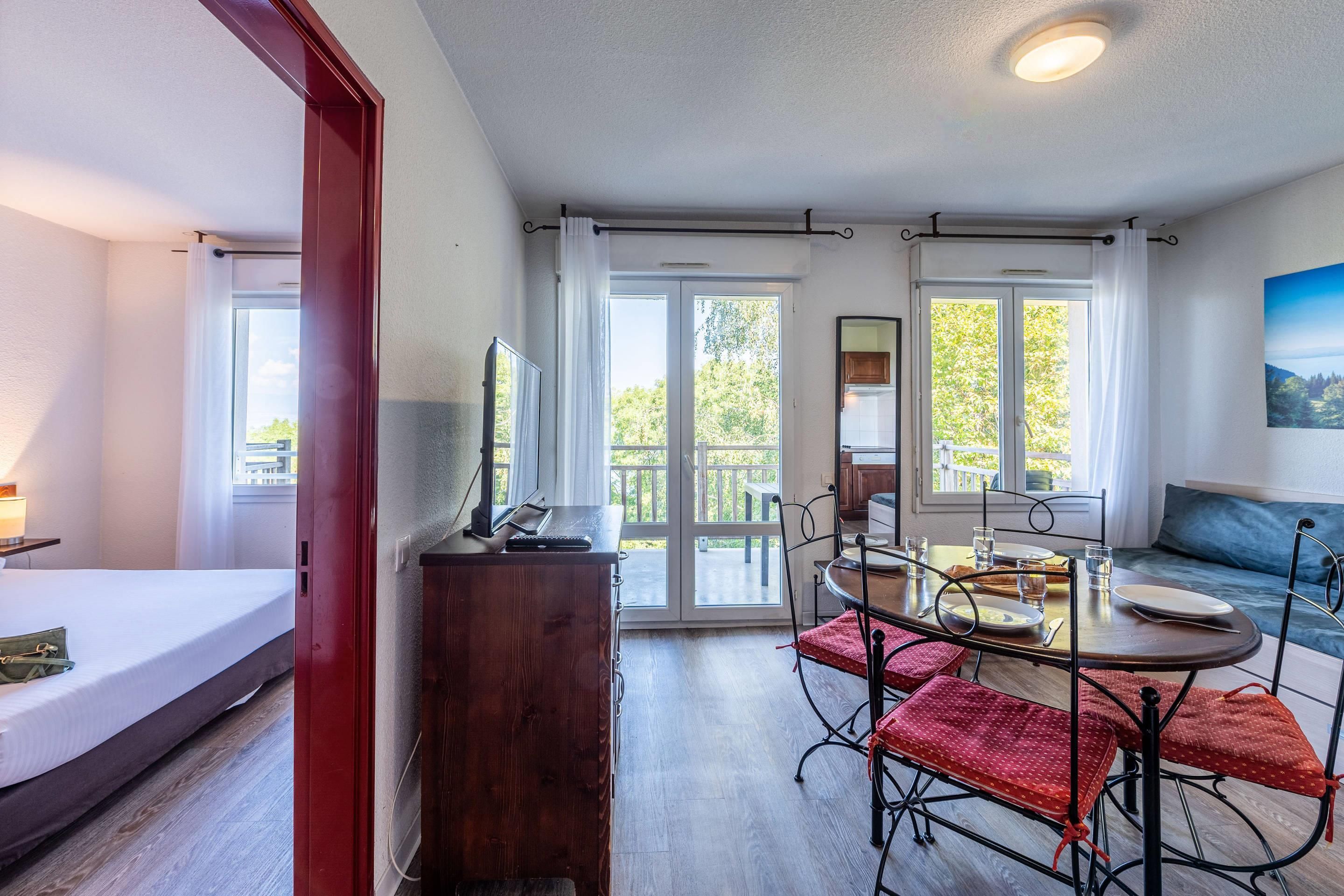 A room with a bed, a desk, a dining table with chairs, and a sofa. There are large windows with curtains, a view of greenery outside, and a painting on the wall.