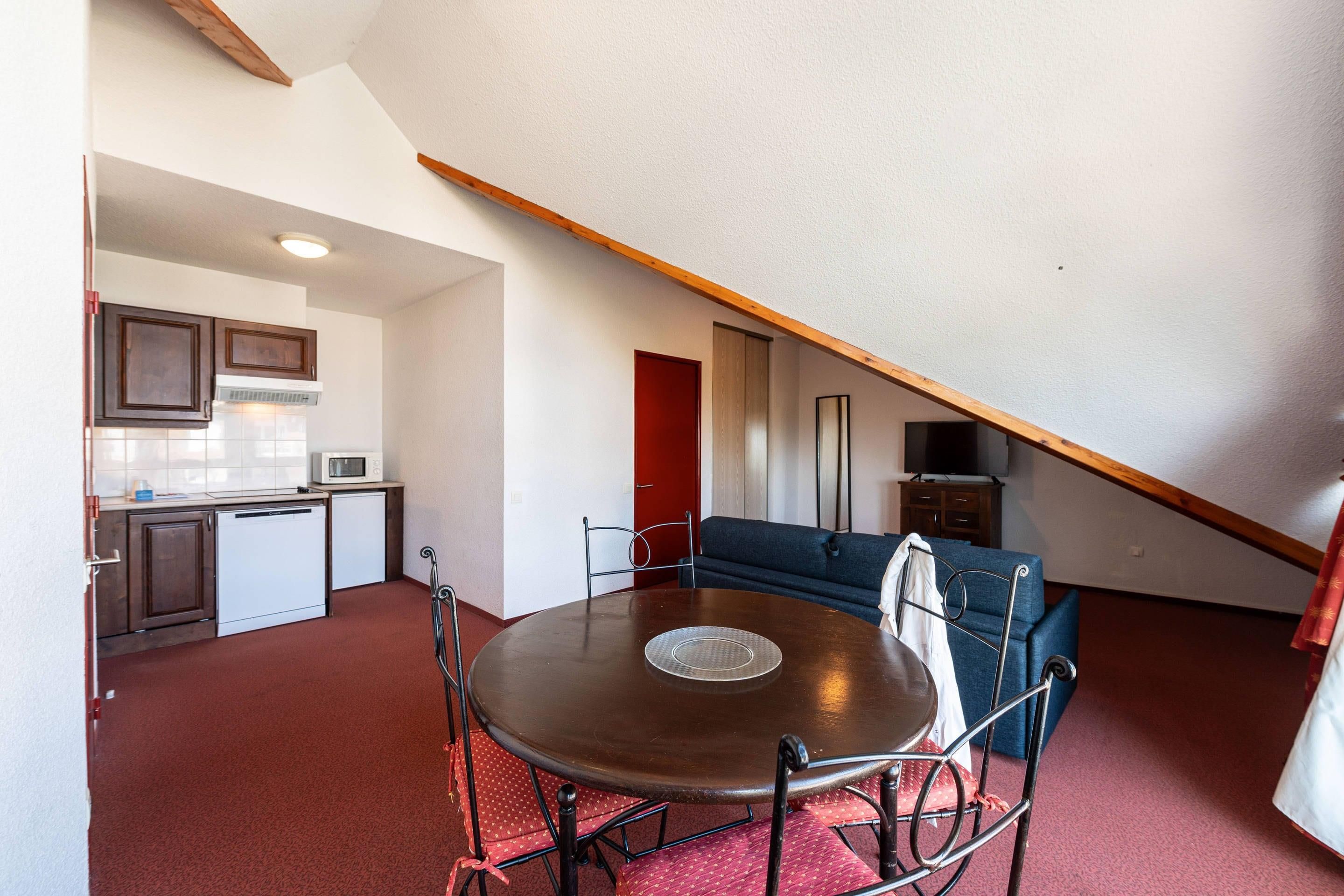 A compact living space with a dining table, kitchen area, and a loft bedroom with a red door.
