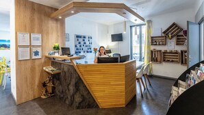 A reception area with a wooden desk, a computer, and a person sitting behind it. There are framed certificates on the wall, a plant, and a shelf with bottles. - Zenitude Evian Les Terrasses du Lac (Evian-les-Bains)