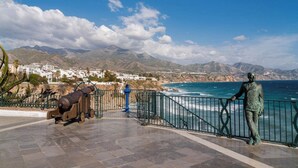 Eine Küstenpromenade mit einer Statue, einer Kanone und Blick auf Meer und Berge.