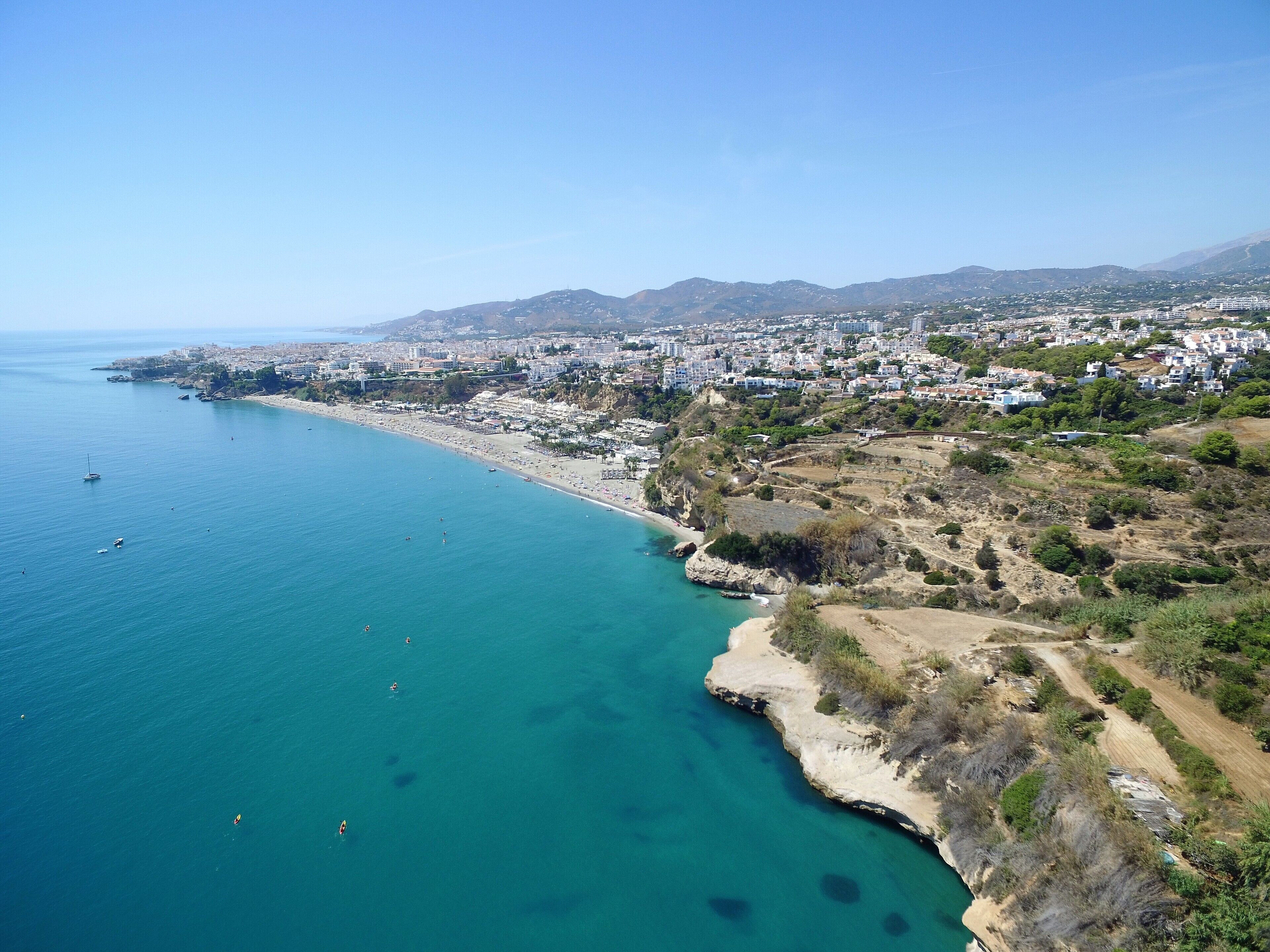 An aerial view of a coastal city with a beach, clear blue waters, and a hilly landscape in the background.