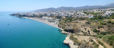 An aerial view of a coastal city with a beach, clear blue waters, and a hilly landscape in the background.